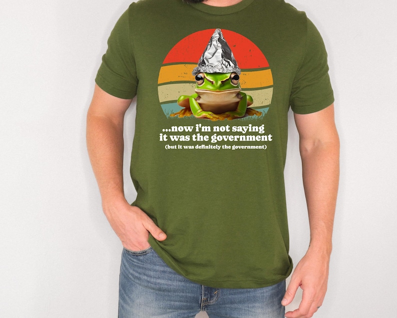 May include: Olive green t-shirt with a cartoon frog wearing a foil hat, set against a retro sunset design. The shirt includes the text: "...now I'm not saying it was the government (but it was definitely the government)".