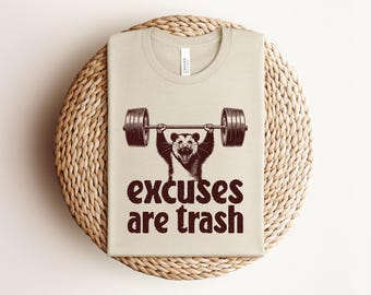 Peut inclure: T-shirt beige avec un opossum brun et blanc soulevant une barre de musculation. Le texte "excuses are trash" est en marron foncé en dessous. Le t-shirt est plié sur un support rond en osier.