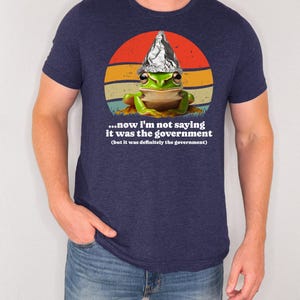 May include: Dark blue t-shirt with a cartoon frog wearing a foil hat, set against a retro sunset design. The text on the shirt reads, "...now I'm not saying it was the government (but it was definitely the government)."