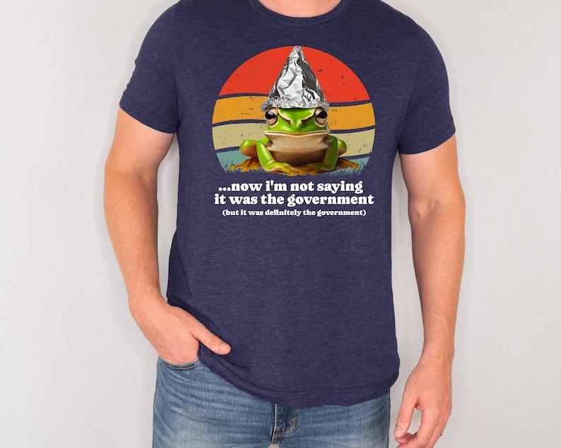 May include: Dark blue t-shirt with a cartoon frog wearing a foil hat, set against a retro sunset design. The text on the shirt reads, "...now I'm not saying it was the government (but it was definitely the government)."