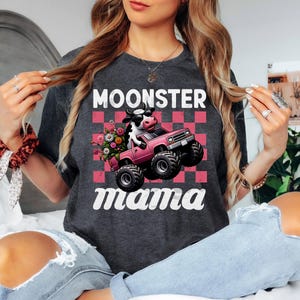 May include: Dark gray t-shirt with the words "MOONSTER Mama" in white. The graphic features a cow driving a pink monster truck with flowers, set against a pink and red checkered background. The shirt is a casual, comfortable style.