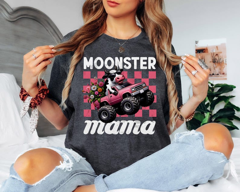 May include: Dark gray t-shirt with the words "MOONSTER Mama" in white. The graphic features a cow driving a pink monster truck with flowers, set against a pink and red checkered background. The shirt is a casual, comfortable style.