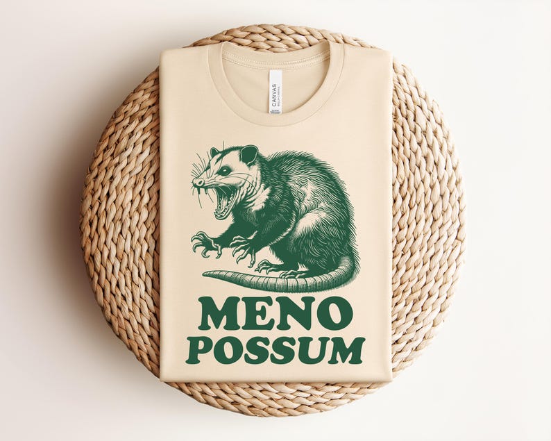 Pu&ograve; includere: Maglietta beige con un'illustrazione verde di un opossum e la scritta "MENO POSSUM". L'opossum &egrave; raffigurato con la bocca aperta e gli artigli. La maglietta &egrave; piegata su un tappetino intrecciato.