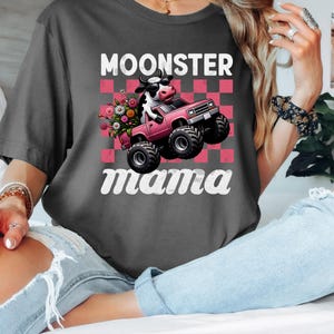 May include: Dark gray t-shirt featuring a cartoon cow driving a pink monster truck with flowers. The text "MOONSTER mama" is printed above and below the graphic in white, with a pink and white checkered background.