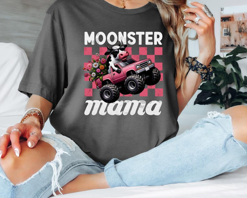 May include: Dark gray t-shirt featuring a cartoon cow driving a pink monster truck with flowers. The text "MOONSTER mama" is printed above and below the graphic in white, with a pink and white checkered background.