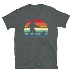K&ouml;nnte beinhalten: Ein dunkel-helles graues T-Shirt mit einer Retro-Sonnenuntergangsgrafik, die eine Silhouette von Bigfoot zeigt, der einen Hund spazieren f&uuml;hrt.