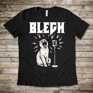 以下が含まれることがあります： 黒いTシャツ。白いイラストで、マイクに向かって歌う猫が描かれています。「BLEGH」の文字が猫の上に太字の白い文字でプリントされています。デザインには、猫とマイクの周りに放射線状の線が含まれています。