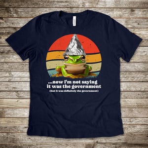 May include: A navy blue t-shirt with a cartoon frog wearing a foil hat, set against a retro sunset design. The text reads "...now I'm not saying it was the government (but it was definitely the government)".