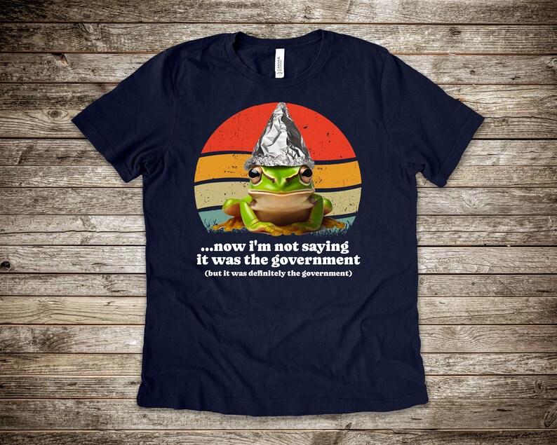May include: A navy blue t-shirt with a cartoon frog wearing a foil hat, set against a retro sunset design. The text reads "...now I'm not saying it was the government (but it was definitely the government)".