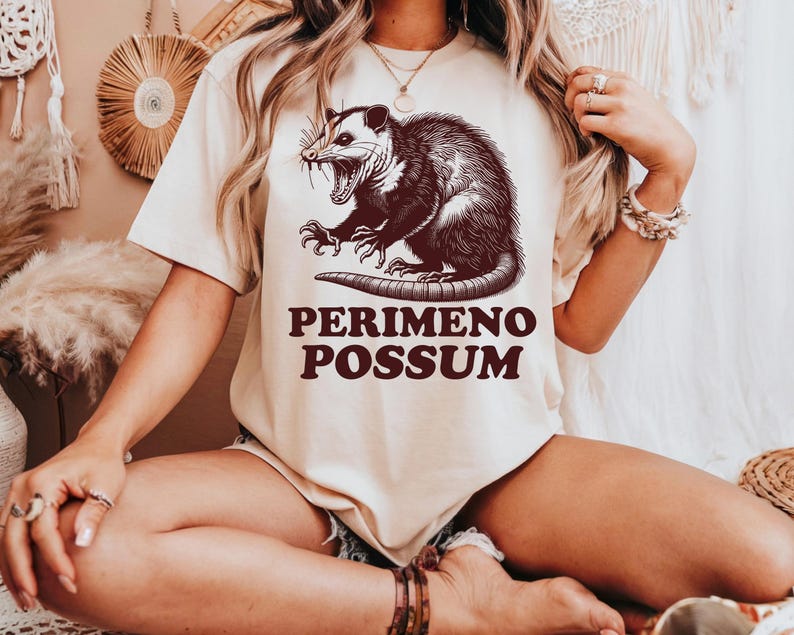 May include: Cream-colored t-shirt featuring a detailed illustration of an opossum with its mouth open, claws extended, and the text "PERIMENO POSSUM" in brown. The shirt is a casual wear item.