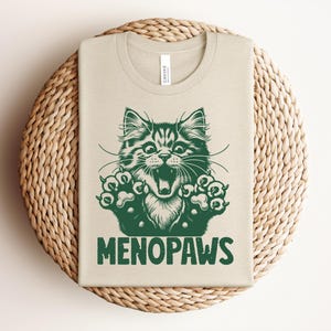 May include: Beige t-shirt with a green illustration of a cat's face and paws, with the word "MENOPAWS" in green block letters. The shirt is folded and placed on a round, woven, beige placemat.