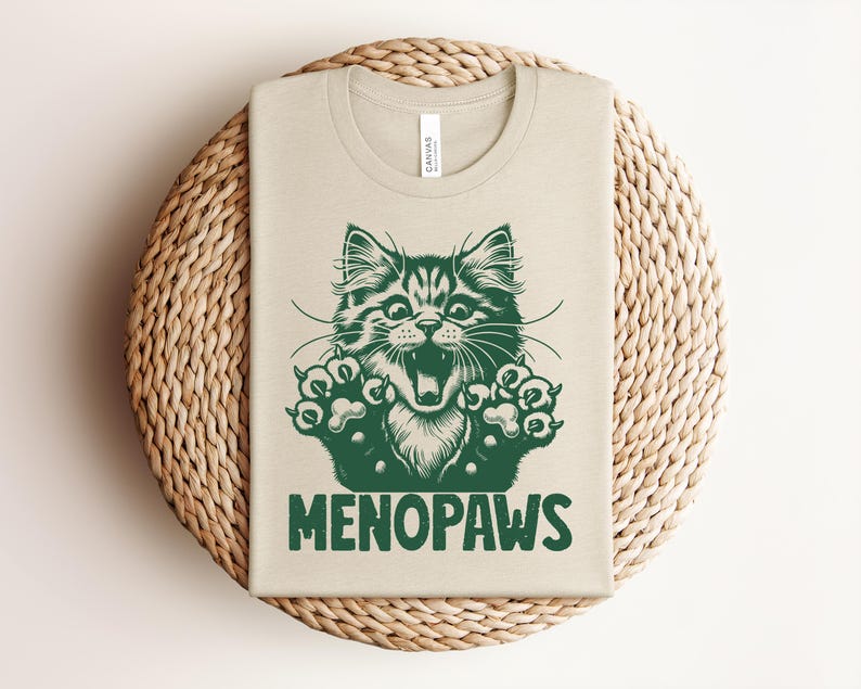 May include: Beige t-shirt with a green illustration of a cat's face and paws, with the word "MENOPAWS" in green block letters. The shirt is folded and placed on a round, woven, beige placemat.