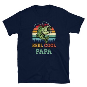 Pu&ograve; includere: Maglietta blu navy con un'illustrazione in stile retr&ograve; di un pesce su un amo da pesca. Il testo "Reel Cool Papa" &egrave; stampato sotto l'immagine.