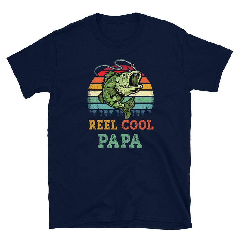 Pu&ograve; includere: Maglietta blu navy con un'illustrazione in stile retr&ograve; di un pesce su un amo da pesca. Il testo "Reel Cool Papa" &egrave; stampato sotto l'immagine.