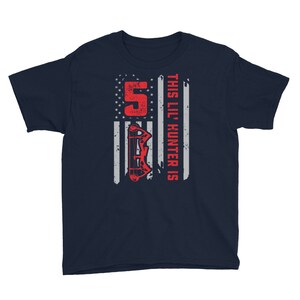 May include: Navy blue youth t-shirt with a red and white American flag design. The flag has a red bow and arrow graphic and the text "This Lil' Hunter is 5".