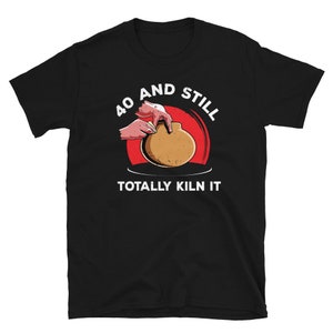 May include: Black t-shirt with a red circle and white text that says "40 and still totally kiln it". Inside the circle is a cartoon hand shaping a clay pot.