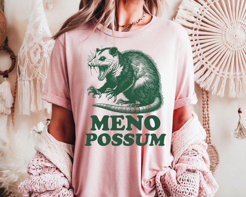 Pu&ograve; includere: T-shirt rosa chiaro con un'illustrazione verde di un opossum e la scritta "MENO POSSUM". L'opossum &egrave; raffigurato con la bocca aperta e gli artigli. La maglietta ha maniche corte.