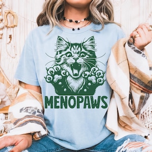 May include: Light blue t-shirt with a green graphic of a cat's face and paws. The cat has an open mouth and appears to be roaring. The word "MENOPAUSE" is printed in green below the cat graphic.