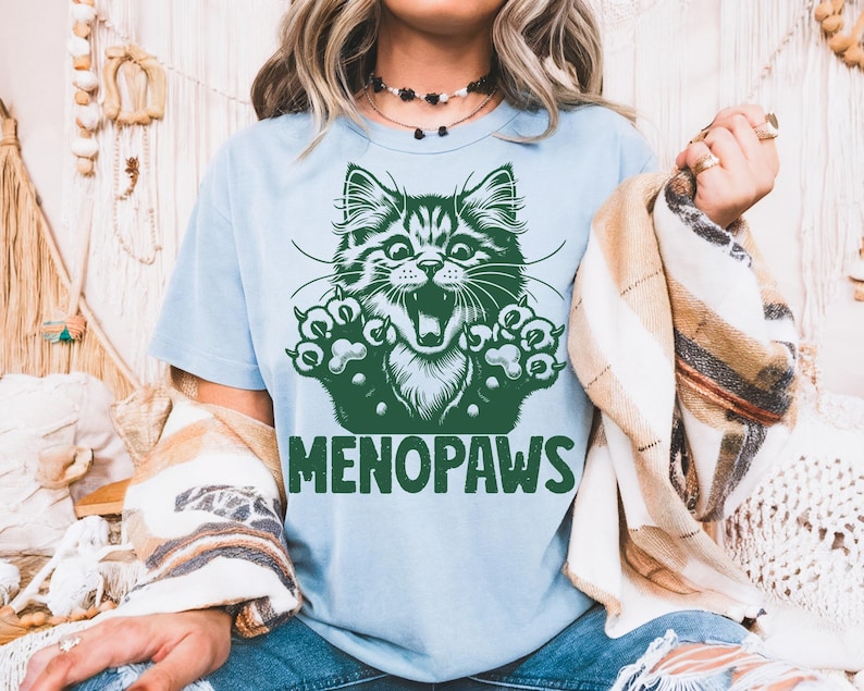 May include: Light blue t-shirt with a green graphic of a cat's face and paws. The cat has an open mouth and appears to be roaring. The word "MENOPAUSE" is printed in green below the cat graphic.