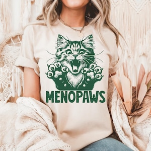 May include: A beige t-shirt featuring a green graphic of a cat with its mouth open and paws outstretched. The text "MENOPAWS" is printed below the cat illustration in green. The shirt is made of a soft material.