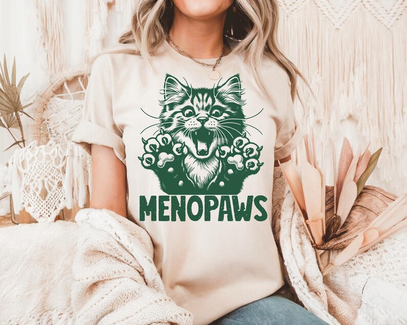 May include: A beige t-shirt featuring a green graphic of a cat with its mouth open and paws outstretched. The text "MENOPAWS" is printed below the cat illustration in green. The shirt is made of a soft material.