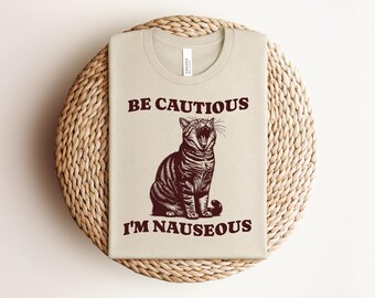 May include: Beige t-shirt with a brown illustration of a yawning cat and the text "BE CAUTIOUS I'M NAUSEOUS". The shirt is folded on a woven, circular surface.