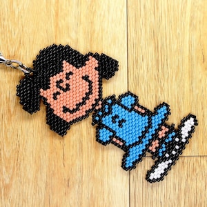 May include: A beaded keychain depicting a smiling face and a snowboarder. The face has black hair and a peach-coloured face. The snowboarder is blue with white and black details. The keychain is attached with a silver clasp. The item is handmade.