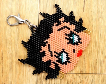Puede incluir: Un llavero de cuentas con un personaje de dibujos animados con cabello negro, piel rosada y ojos azules. El llavero tiene un cierre de metal plateado.