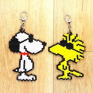 Op de afbeelding: Twee kralen sleutelhangers met cartoonfiguren. De eerste sleutelhanger is een witte en zwarte kralen Snoopy, met een rode kraag. De tweede sleutelhanger is een gele en zwarte kralen Woodstock.
