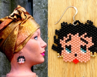 May include: A mannequin head with a gold-toned head wrap and beaded earrings. The earrings depict a cartoon face with black hair, pink skin, blue eyes, and a red mouth. The earrings are on a silver hook.