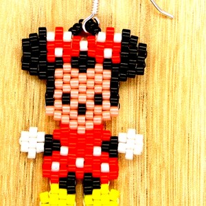 Peut inclure: Une paire de boucles d'oreilles en perles repr&eacute;sentant un dessin pixelis&eacute; de Minnie Mouse. Les boucles d'oreilles sont rouges, noires, blanches et jaunes. Les boucles d'oreilles sont attach&eacute;es &agrave; des crochets argent&eacute;s.
