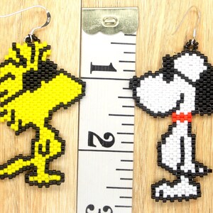 May include: A pair of beaded earrings featuring the characters Snoopy and Woodstock from the Peanuts comic strip. The Snoopy earring is white with black details and a red collar. The Woodstock earring is yellow with black details. The earrings are approximately 5 cm long.