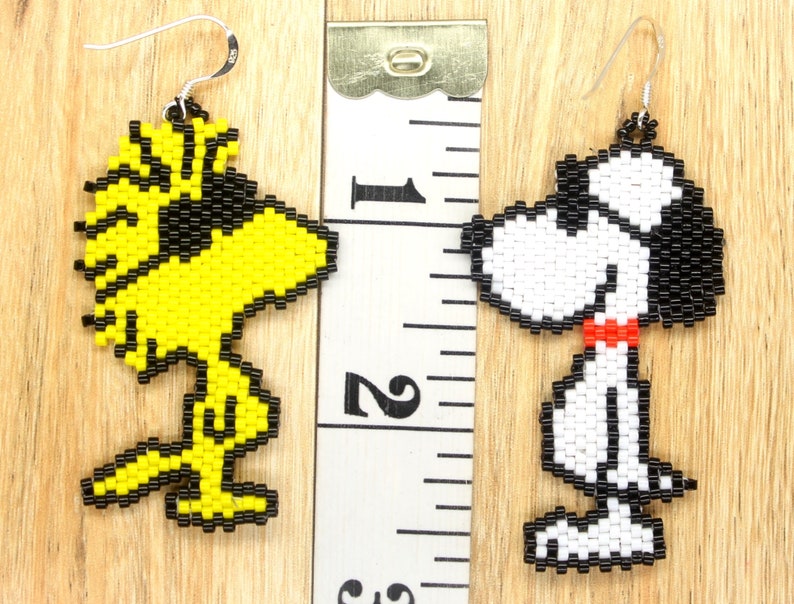 May include: A pair of beaded earrings featuring the characters Snoopy and Woodstock from the Peanuts comic strip. The Snoopy earring is white with black details and a red collar. The Woodstock earring is yellow with black details. The earrings are approximately 5 cm long.