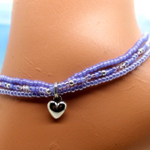 May include: A delicate, beaded anklet in shades of lavender and silver. The anklet features three strands of small, round beads and a small, silver heart charm. The jewelry is designed to be worn around the ankle.