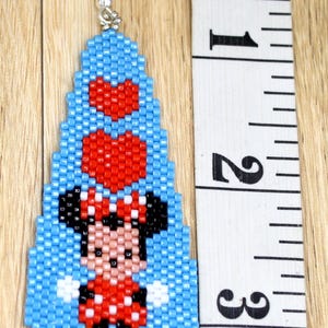 May include: Handmade beaded earrings with a cartoon character design. The earrings are approximately 6.35 cm long. The design includes a character in a red dress with a white polka dot pattern, black ears, and a red bow. The background is light blue.