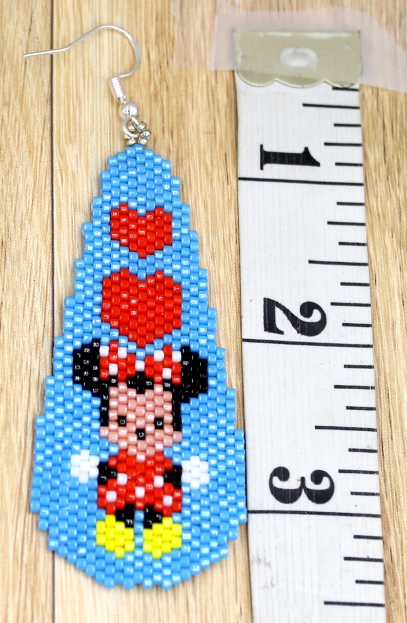 May include: Handmade beaded earrings with a cartoon character design. The earrings are approximately 6.35 cm long. The design includes a character in a red dress with a white polka dot pattern, black ears, and a red bow. The background is light blue.