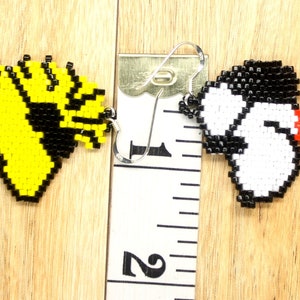 May include: A pair of beaded earrings featuring cartoon characters. The left earring is yellow and black and depicts a bird. The right earring is white and black and depicts a dog. The earrings are approximately 2.5 cm in length.