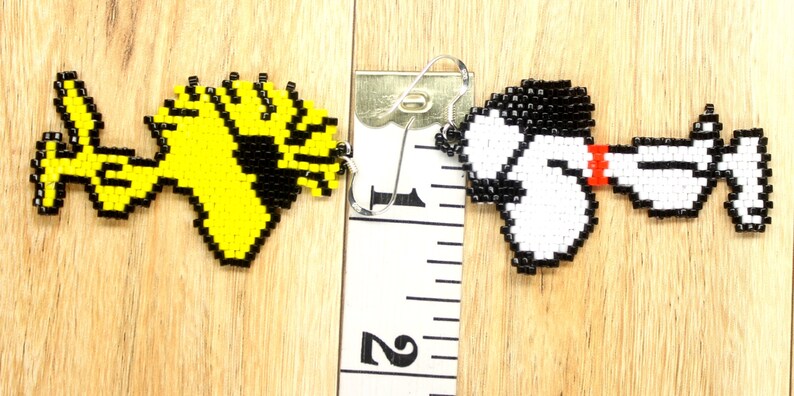 May include: A pair of beaded earrings featuring cartoon characters. The left earring is yellow and black and depicts a bird. The right earring is white and black and depicts a dog. The earrings are approximately 2.5 cm in length.