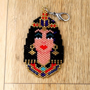 May include: A beaded keychain featuring a colorful depiction of an Egyptian queen with a crown, black hair, and a turquoise jewel. The keychain has a silver metal clasp.