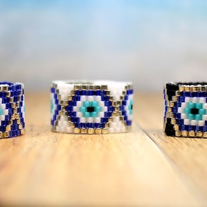 May include: Three beaded rings with a blue evil eye design. The rings are on a wooden surface. The rings are blue, white, and black.