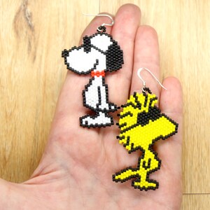 May include: A pair of beaded earrings featuring cartoon characters Snoopy and Woodstock. Snoopy is white with black eyes and a red collar. Woodstock is yellow with black eyes. The earrings are hanging from silver hooks.