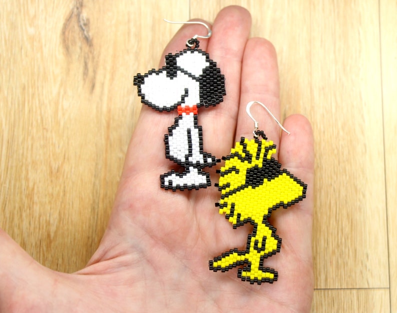 May include: A pair of beaded earrings featuring cartoon characters Snoopy and Woodstock. Snoopy is white with black eyes and a red collar. Woodstock is yellow with black eyes. The earrings are hanging from silver hooks.