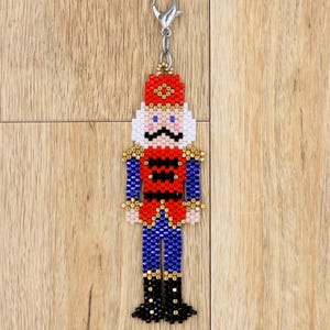 Peut inclure: Un charm de casse-noisette perlé en rouge, bleu, noir et or. Le charm présente un chapeau rouge, une moustache noire et des yeux bleus. Le charm est attaché à un fermoir argenté.