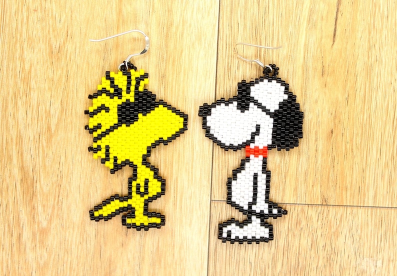 May include: A pair of beaded earrings featuring cartoon characters. The left earring is yellow and black and depicts the character Woodstock from the Peanuts comic strip. The right earring is white, black, and red and depicts the character Snoopy from the Peanuts comic strip.