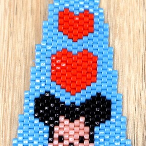 May include: A beaded earring with a cartoon character design. The earring is mainly light blue with red heart accents and a character face in black, pink, and white. It has a silver-coloured hook.