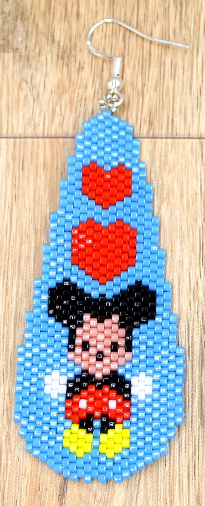 May include: A beaded earring with a cartoon character design. The earring is mainly light blue with red heart accents and a character face in black, pink, and white. It has a silver-coloured hook.