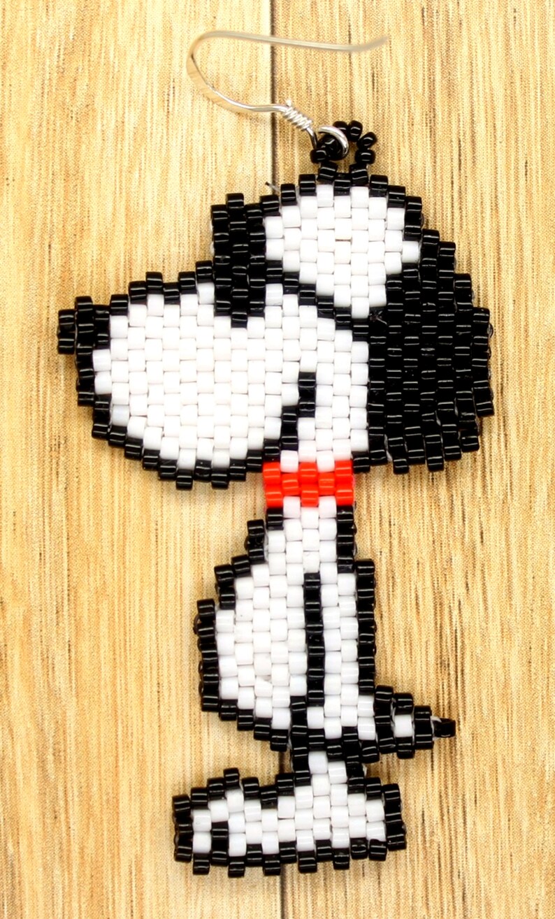 May include: Beaded earrings featuring a black and white cartoon dog with a red bow tie. The earrings are made with small, round beads and have a silver hook.