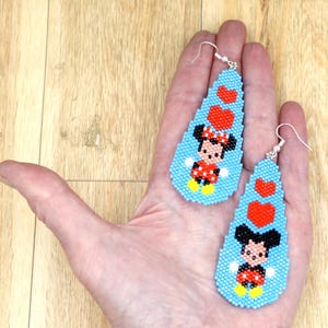 May include: Handmade beaded earrings with a cartoon character design. The earrings are light blue, teardrop-shaped, and decorated with red hearts and a cartoon character in a red and black outfit. They have silver-coloured hooks.