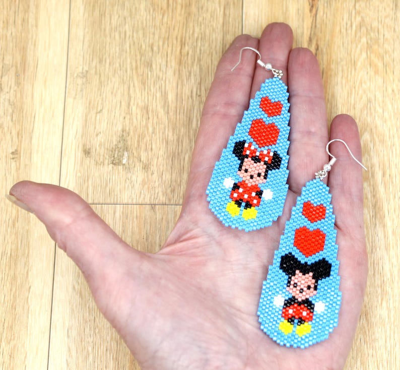 May include: Handmade beaded earrings with a cartoon character design. The earrings are light blue, teardrop-shaped, and decorated with red hearts and a cartoon character in a red and black outfit. They have silver-coloured hooks.