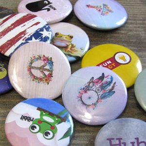 May include: Assortment of colorful round buttons with various designs, including a peace sign, a frog, an American flag, a lightbulb, and a dreamcatcher. The buttons are all approximately 2 inches in diameter.
